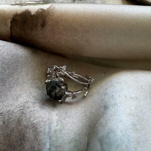 May include: A silver ring with a dark gray stone set in a branch-like design. The ring is on a white fabric background.