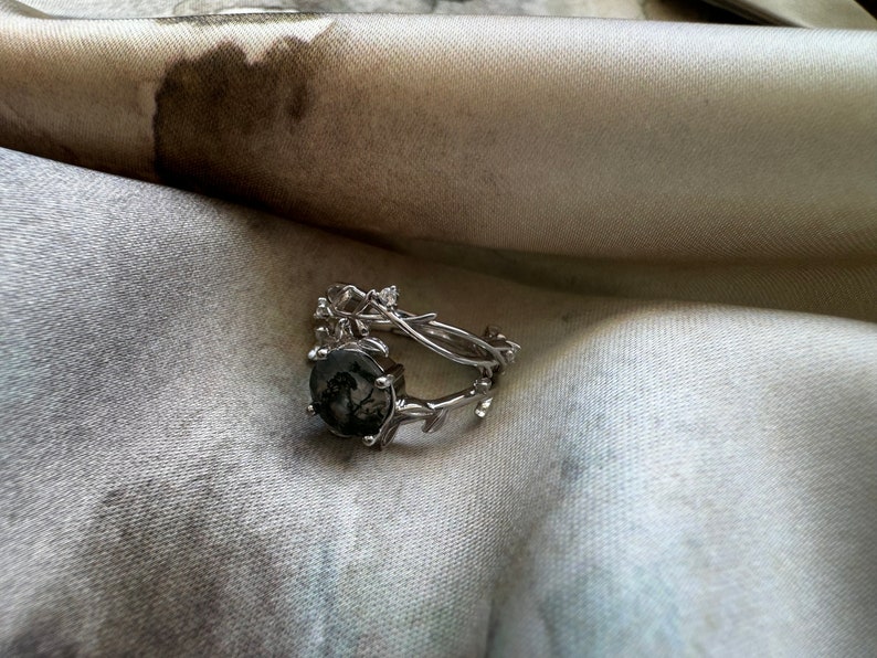 May include: A silver ring with a dark gray stone set in a branch-like design. The ring is on a white fabric background.