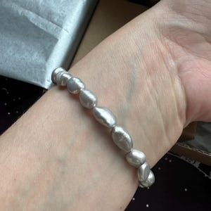 May include: A silver pearl bracelet with oval-shaped pearls. The bracelet is on a person's wrist.