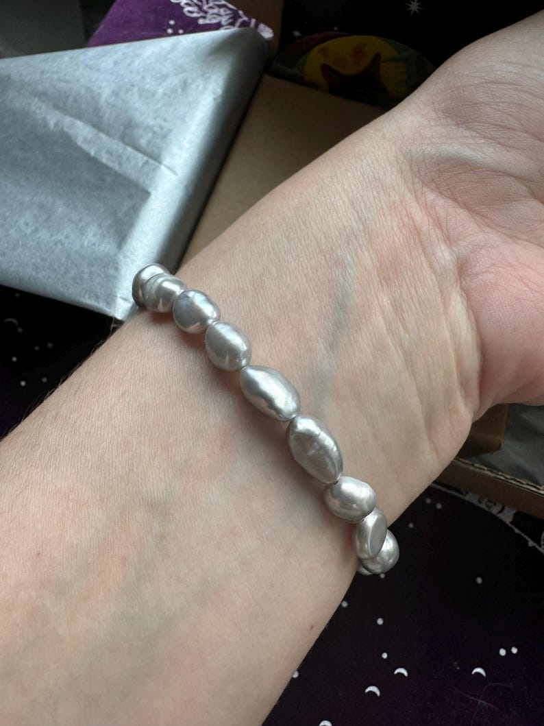 May include: A silver pearl bracelet with oval-shaped pearls. The bracelet is on a person's wrist.