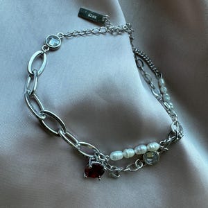 May include: A silver chain necklace with multiple layers, featuring a red gemstone pendant, white pearls, and clear crystal accents. The necklace has a silver tag with the text "925" on it.