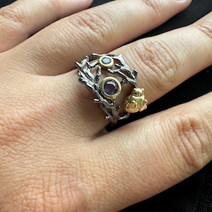 May include: A silver ring with a branch design featuring three gemstones. The ring has a purple gemstone, a yellow gemstone, and a gold-colored heart charm.