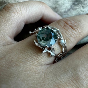 May include: A silver ring featuring a green moss agate gemstone set in a delicate branch design. The ring has a double band with a smaller diamond accent.
