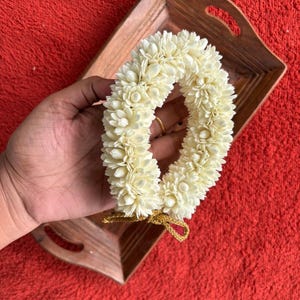 May include: A cream-colored floral garland, likely jasmine, held in a hand. The garland is arranged in a circular shape and tied with a gold-colored string. The background is a wooden tray on a red surface.