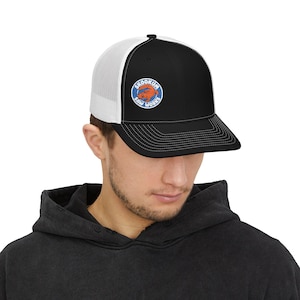 May include: Black and white trucker hat with a curved brim. The front panel is black, and the back is white mesh. A circular patch on the front displays the text "SKOOKUM SAW WORKS" around a cartoon beaver in orange and blue.