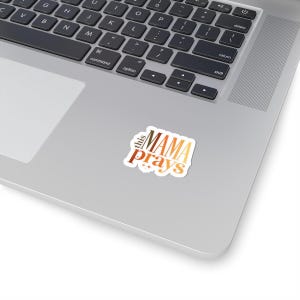 May include: A sticker with the text "this MAMA prays" in a gradient of brown and gold colors. The sticker is on a laptop, with the keyboard visible in the background.