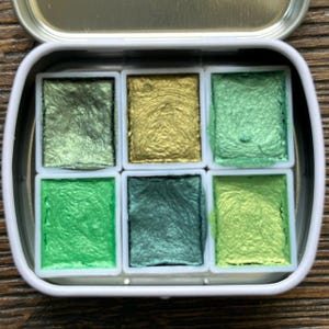 May include: A metal tin containing six rectangular pans of metallic watercolor paint. The colors include various shades of green, ranging from dark olive and forest green to bright lime and a shimmering gold, all with a textured, pearlescent finish.