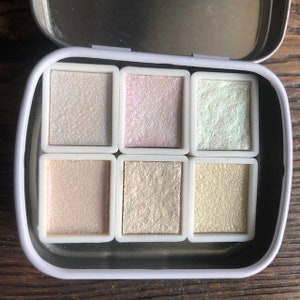 Op de afbeelding: Een blik met zes iriserende aquarelverf in verschillende tinten, waaronder wit, roze, groen en goud.