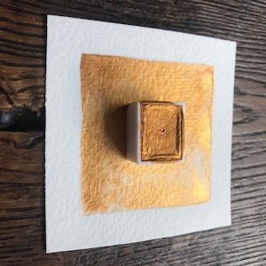 May include: A square of white watercolor paper with a gold metallic paint swatch and a small, rectangular paint pan. The gold paint is applied in a square shape on the paper, and the paint pan is white with a gold paint square.