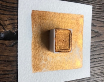 May include: A square of white watercolor paper with a gold metallic paint swatch and a small, rectangular paint pan. The gold paint is applied in a square shape on the paper, and the paint pan is white with a gold paint square.