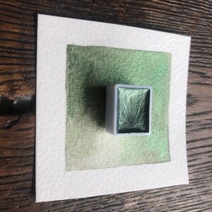 May include: A square of watercolor paper with a green wash, and a small, rectangular watercolor paint pan. The paint pan is filled with a shimmering, dark green watercolor paint.