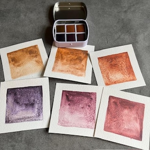 May include: A white metal watercolor paint set with six colors, open to reveal the paints. Six square watercolor swatches in shades of brown, orange, and purple are arranged around the paint set on a gray surface. The swatches are on white paper.