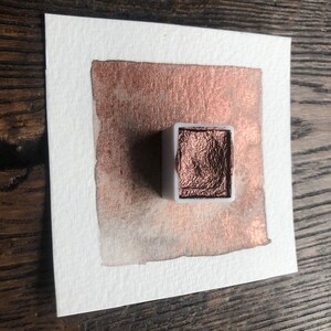 May include: A square of watercolor paper with a copper-colored metallic paint swatch. A small, white plastic container of the same metallic paint sits on top of the paper. The paper is on a wooden surface.