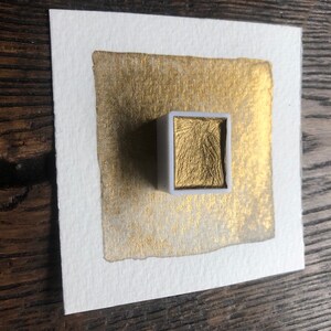 May include: A square watercolor painting on white paper with a gold metallic wash. A small, white, rectangular watercolor pan filled with gold paint sits on top of the painting. The paper is on a wooden surface.