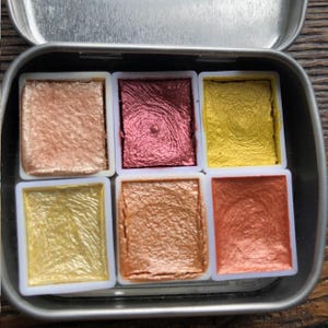 May include: A metal tin containing six metallic watercolor paints in shades of gold, copper, and rose. The paints are in individual white plastic containers, arranged in two rows of three. The tin is open, revealing the vibrant colors.