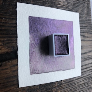 May include: A small, square pan of metallic purple watercolor paint. The pan is sitting on a white sheet of paper with a sample of the paint applied to the paper.