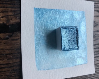 May include: A small square watercolor paint pan with a metallic blue pigment. The paint is on a white paper surface and has a shimmery finish.