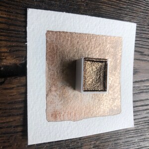 May include: A square of watercolor paper with a copper-colored paint swatch and a small, white watercolor paint pan filled with metallic gold paint. The paper is set on a wooden surface.