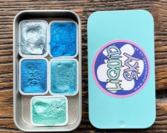 May include: A set of five metallic watercolour paints in a silver tin. The paints are in shades of blue and teal. The tin has a light blue lid with a circular logo that says "Liquid Sky" in a stylized font.