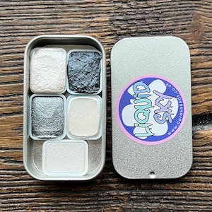May include: A silver metal watercolor paint palette with five metallic shades. The open palette reveals a range of colors, including white, black, and silver. The lid features a Liquid Sky logo.