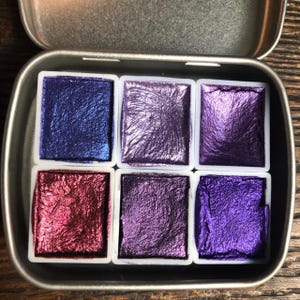 May include: A metal tin with six watercolor paint pans. The pans are arranged in two rows of three. The colors are a deep blue, a light purple, a dark purple, a red, a light purple, and a dark purple.