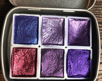 May include: A metal tin with six watercolor paint pans. The pans are arranged in two rows of three. The colors are a deep blue, a light purple, a dark purple, a red, a light purple, and a dark purple.
