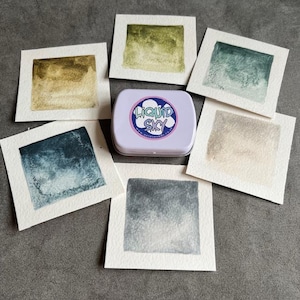 May include: A watercolour paint set featuring various colours, including greens, blues, and browns, presented on individual white cards. A small, lilac tin with the text "LIQUID SKY" is in the centre.
