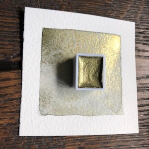 May include: A square of watercolor paper with a gold metallic paint swatch and a small, square, gold watercolor paint pan. The paper has a gold wash in the center and a white border. The paint pan is light gray.