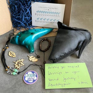 May include: A teal ceramic 12-hole ocarina, a black carrying case, a fingering chart, a wooden bead bracelet, two wooden leaf charms, a black cord necklace with light green beads, a circular sticker, and a handwritten note on green paper.