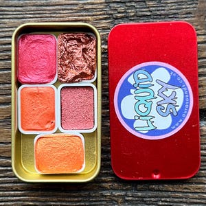 May include: A gold metal watercolor paint palette with six vibrant colors. The colors include shades of red, orange, and copper. The red lid of the palette has a sticker with the text "Liquid Sky".