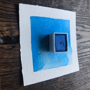 May include: A small, white, square watercolor pan with a blue watercolor paint inside. The pan is sitting on a white sheet of paper with a blue watercolor wash.