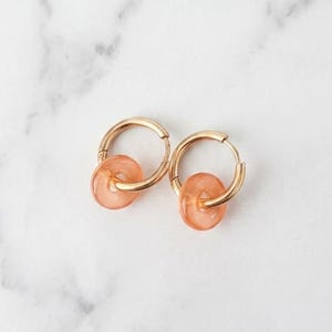 May include: A pair of gold hoop earrings with a small, peach-colored, circular bead dangling from each hoop. The earrings are displayed on a white and gray marble surface.