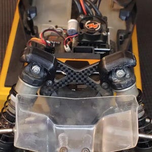 May include: A close-up of a black carbon fiber shock tower with two black shock absorbers attached. The shock tower has a checkered pattern and is mounted on a yellow and black vehicle.
