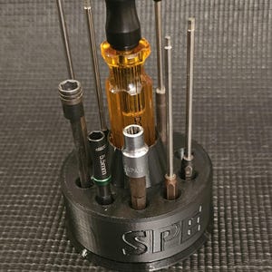 May include: A black plastic screwdriver holder with a circular base and eight slots for holding screwdrivers. The holder has the word "SPE" printed on the side. There are seven screwdrivers in the holder, including a yellow and black screwdriver with a black handle.