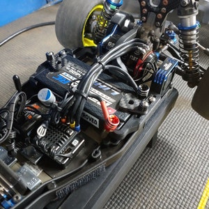 May include: A black and blue remote control car chassis with a black battery pack, a black and silver electronic speed control, and a red and black wire. The car is on a gray carpet.