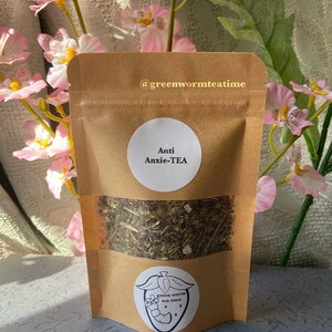 Puede incluir: Una bolsa de papel marr&oacute;n con una etiqueta blanca que dice "Anti Anxie-TEA" y un logotipo de gusano verde. La bolsa contiene t&eacute; de hojas sueltas.