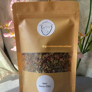 Puede incluir: Una bolsa de papel marr&oacute;n con una ventana transparente que muestra una mezcla de hierbas y flores secas. La bolsa tiene una pegatina blanca con el texto "green worm tea time" y un gusano de dibujos animados dentro de una fresa. La bolsa tambi&eacute;n tiene una pegatina blanca con el texto "Anti Anxie-TEA".