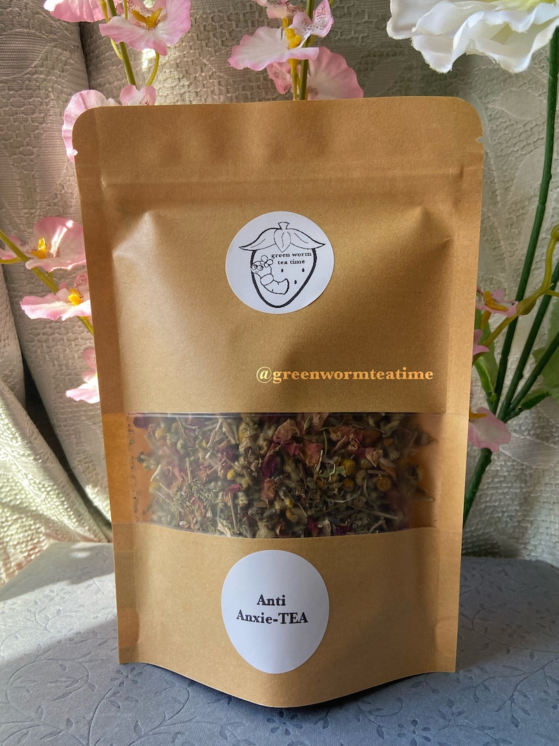 Puede incluir: Una bolsa de papel marr&oacute;n con una ventana transparente que muestra una mezcla de hierbas y flores secas. La bolsa tiene una pegatina blanca con el texto "green worm tea time" y un gusano de dibujos animados dentro de una fresa. La bolsa tambi&eacute;n tiene una pegatina blanca con el texto "Anti Anxie-TEA".