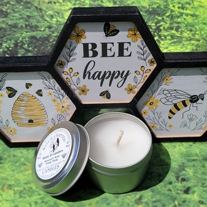 Può includere: Una piccola candela in latta con cera bianca e uno stoppino di legno. La candela è in una latta argentata con un'etichetta in bianco e nero che dice "Witch Crafted Candles" e "Honey & Hive" con un'illustrazione di un'ape. La candela è davanti a un cartello esagonale di legno con il testo "BEE happy" e illustrazioni di api.