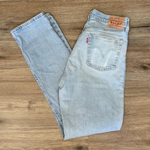 Könnte beinhalten: Hellblaue Jeans aus Denim mit klassischem Fünf-Taschen-Design. Die Jeans haben ein rotes Levi's-Etikett auf der Gesäßtasche und einen braunen Lederaufnäher. Die Jeans liegen flach auf einer Holzoberfläche.