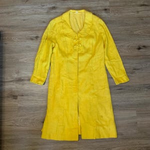 May include: A bright yellow long-sleeved coat featuring a rounded Peter Pan collar and two decorative fabric-covered toggle closures at the neckline. The coat has a structured fit with vertical seam detailing and a straight hemline.