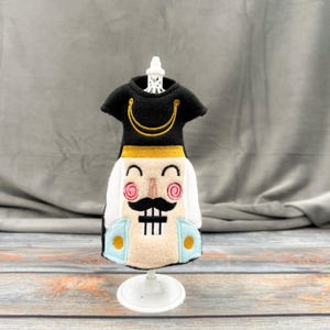 May include: A decorative doll dress shaped like a nutcracker soldier. The black top has a gold trim, and the face is beige with a black mustache and pink cheeks. The doll dress is on a white stand.