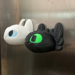 May include: Two 3D printed dragon figurines, one black with a green eye and one white with a blue eye, are attached to a silver surface.