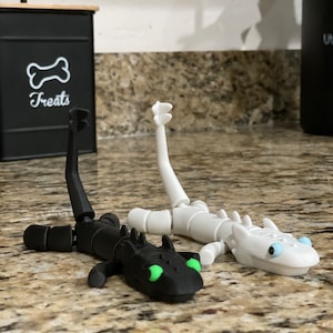 May include: Two articulated dragon figurines, one black with green eyes and one white with blue eyes. The dragons are posed in a playful manner, with their tails curled up.
