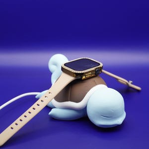 May include: A gold Apple Watch with a diamond-encrusted bezel is resting on a blue and brown Squirtle figurine. The watch is attached to a beige band and is plugged into a charging cable.