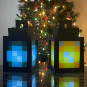 May include: Two black Minecraft-themed lanterns with light blue and yellow pixelated designs. The lanterns are illuminated and sitting on a dark surface in front of a Christmas tree with lights.