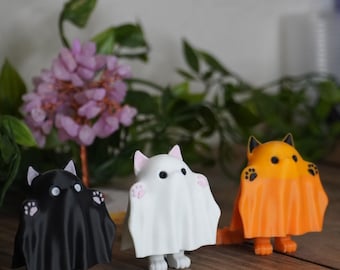 May include: Three small cat figurines dressed as ghosts for Halloween. The cats are black, white, and orange. They are standing on a wooden surface.