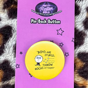 May include: A yellow pin-back button with the text "BOYS ARE STUPID, THROW ROCKS AT THEM!" and a cartoon illustration. The button is on a purple card with the text "Pin-Back Button" and the brand name "Pretty Rotten Girls".
