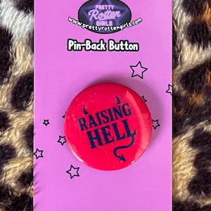 May include: A red pin-back button with the words "RAISING HELL" in dark blue, with a devil tail graphic. The button is on a pink card with the text "Pin-Back Button" and the Pretty Rotten Girls logo.