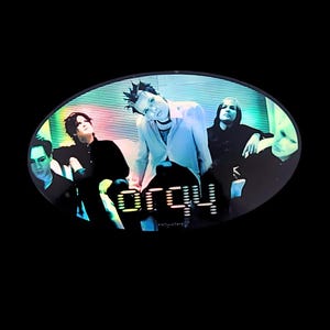 May include: An oval image featuring a band with the word "Orgy" in a stylized font. The band members are dressed in dark clothing, with a mix of black and light-colored shirts. The background has a striped pattern.